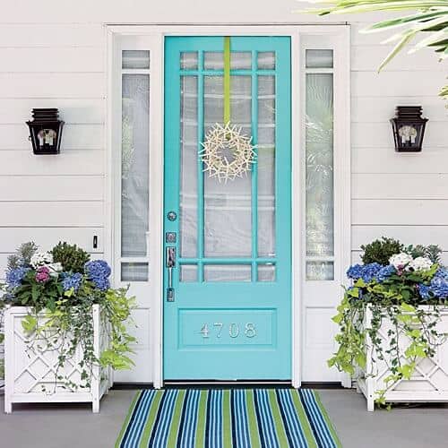 Front Porch with turquoise door, seashell wreath, white resin planter boxes, blue and green striped front door mat indoor outdoor rug